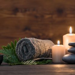 Brown towel on green fern with candles and black hot stone on wooden background. Hot stone massage setting lit by candles. Hot stone therapy for one person with candle light. Beauty spa treatment and relax concept.